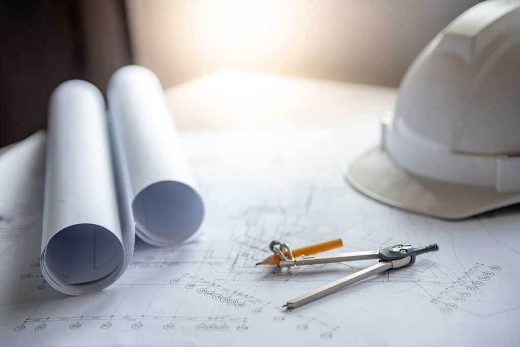 Architectural blueprints with rolled plans, drafting compass, pencil, and a white hard hat on a work surface.