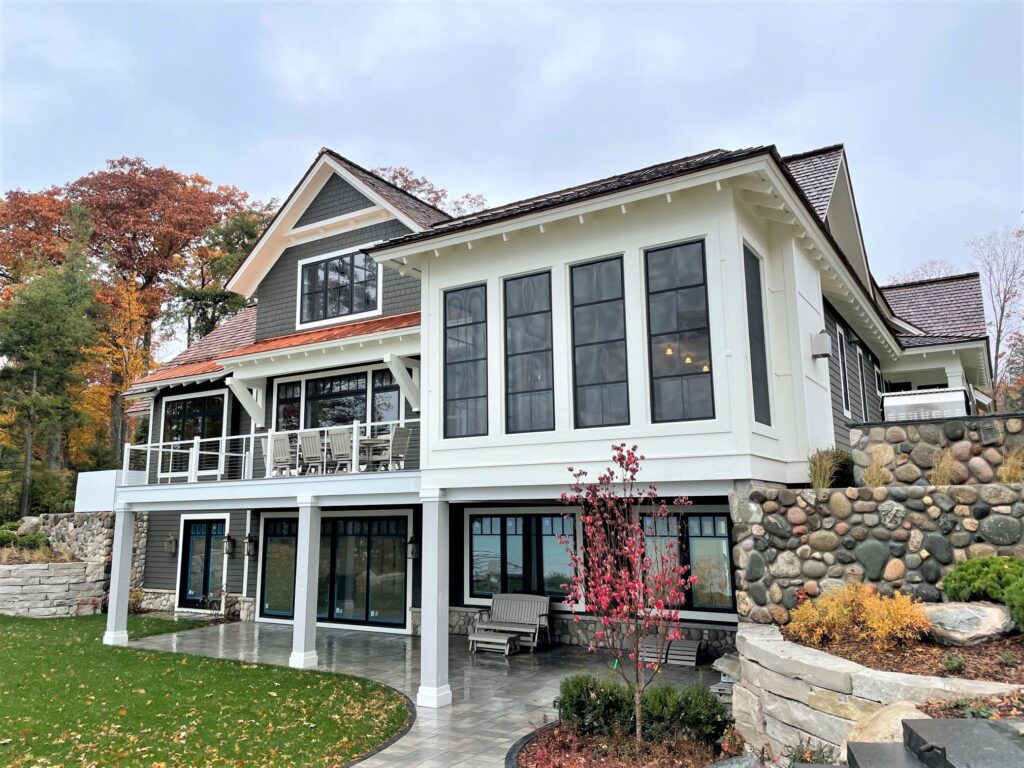 Large custom lakefront home with covered patio, elevated deck, and stone retaining walls set into a landscaped hillside.