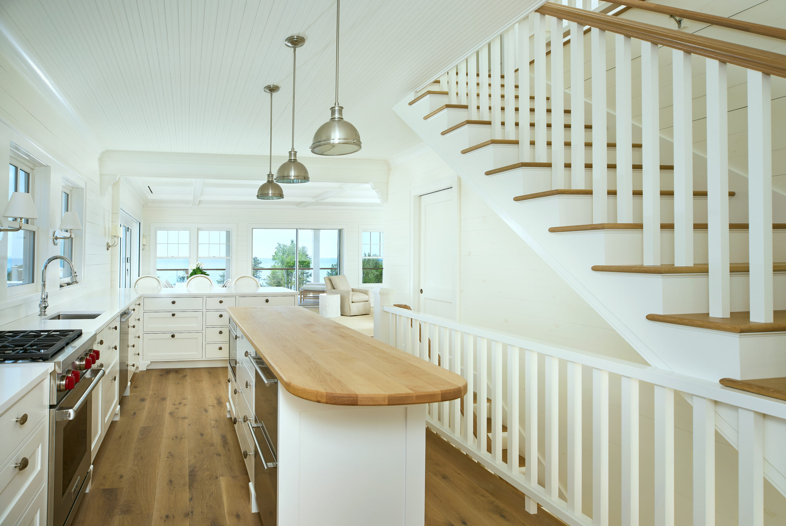 White kitchen with butcher block island and staircase featuring natural wood treads and white spindles, with a view of Lake Michigan through large windows.