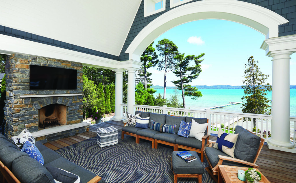 Covered lakefront porch with stone fireplace, outdoor seating, and arched opening framing a view of the water and dock.
