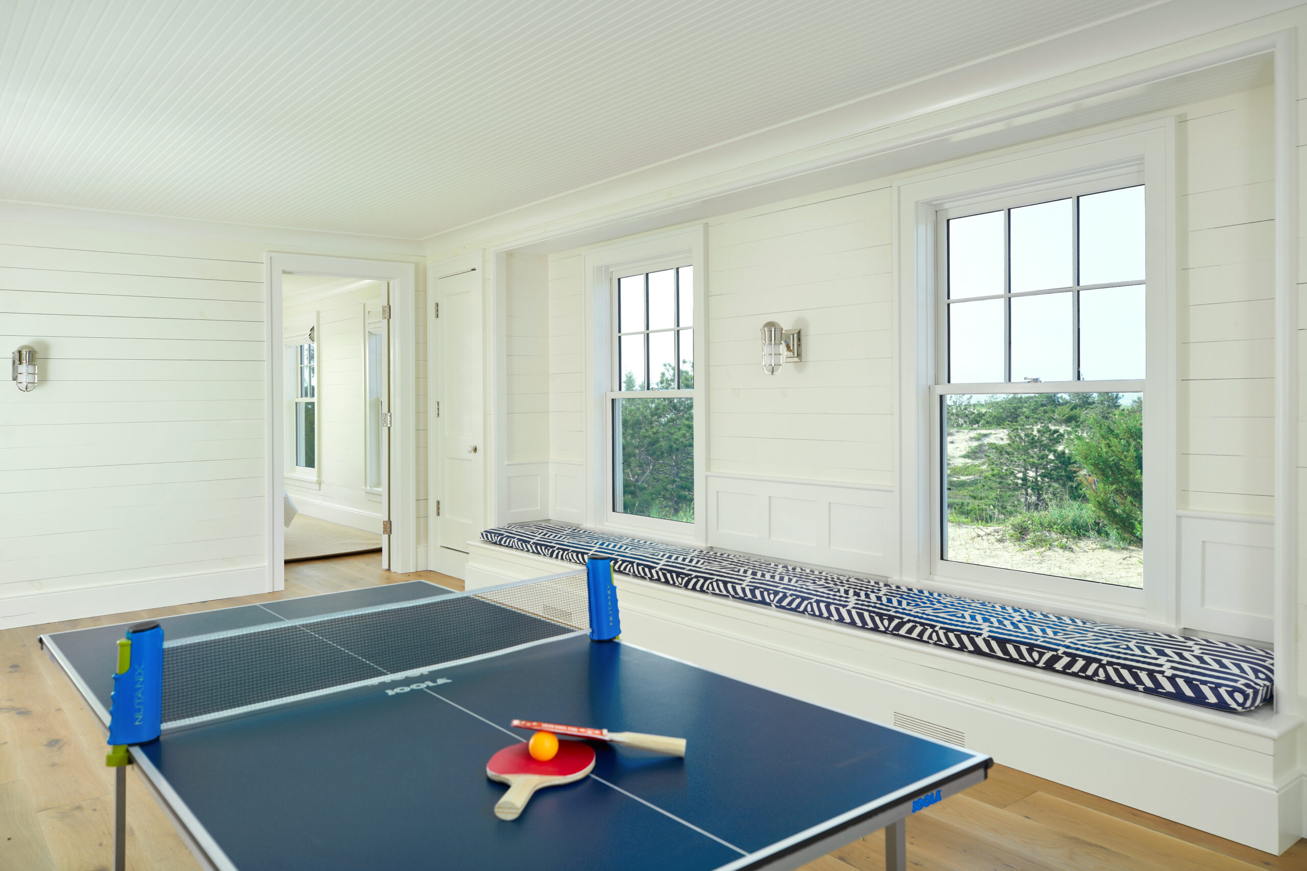 Ping pong table with paddles in a bright white shiplap game room featuring built-in window seating and large windows overlooking sand dunes and greenery.