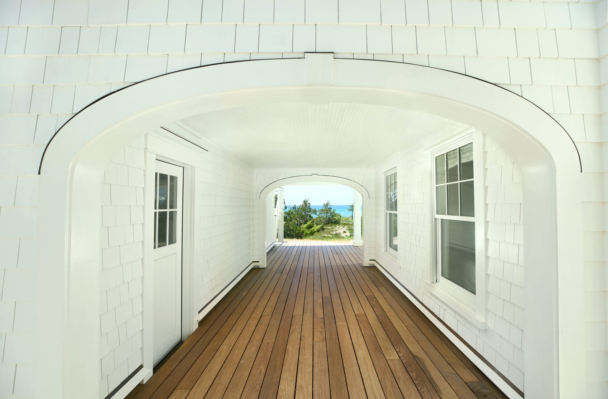 White shingled covered walkway with wood flooring and arched openings on both ends, offering a view of Lake Michigan through the far arch.