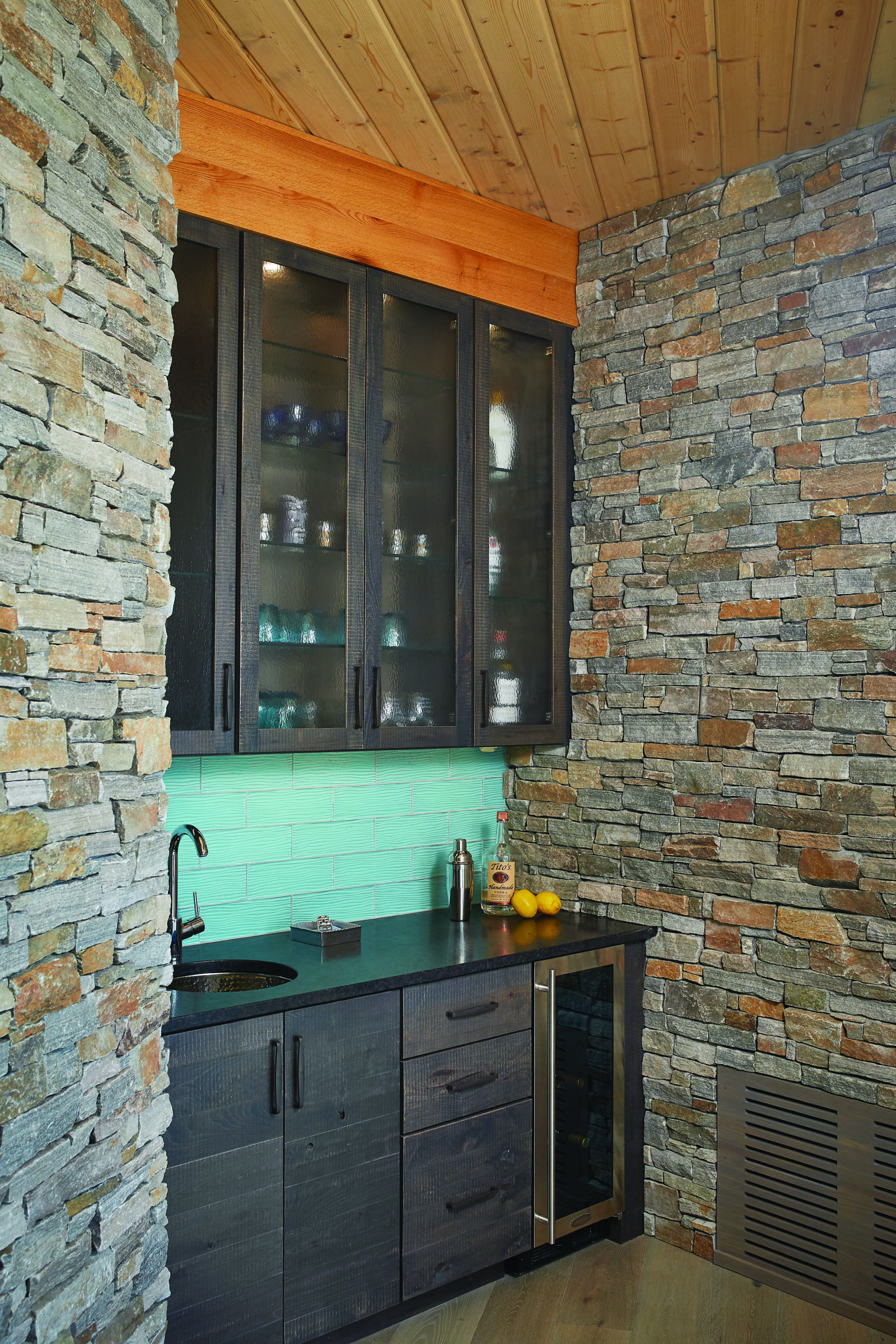 Custom home bar with stone walls, glass-front cabinets, and turquoise tile backsplash