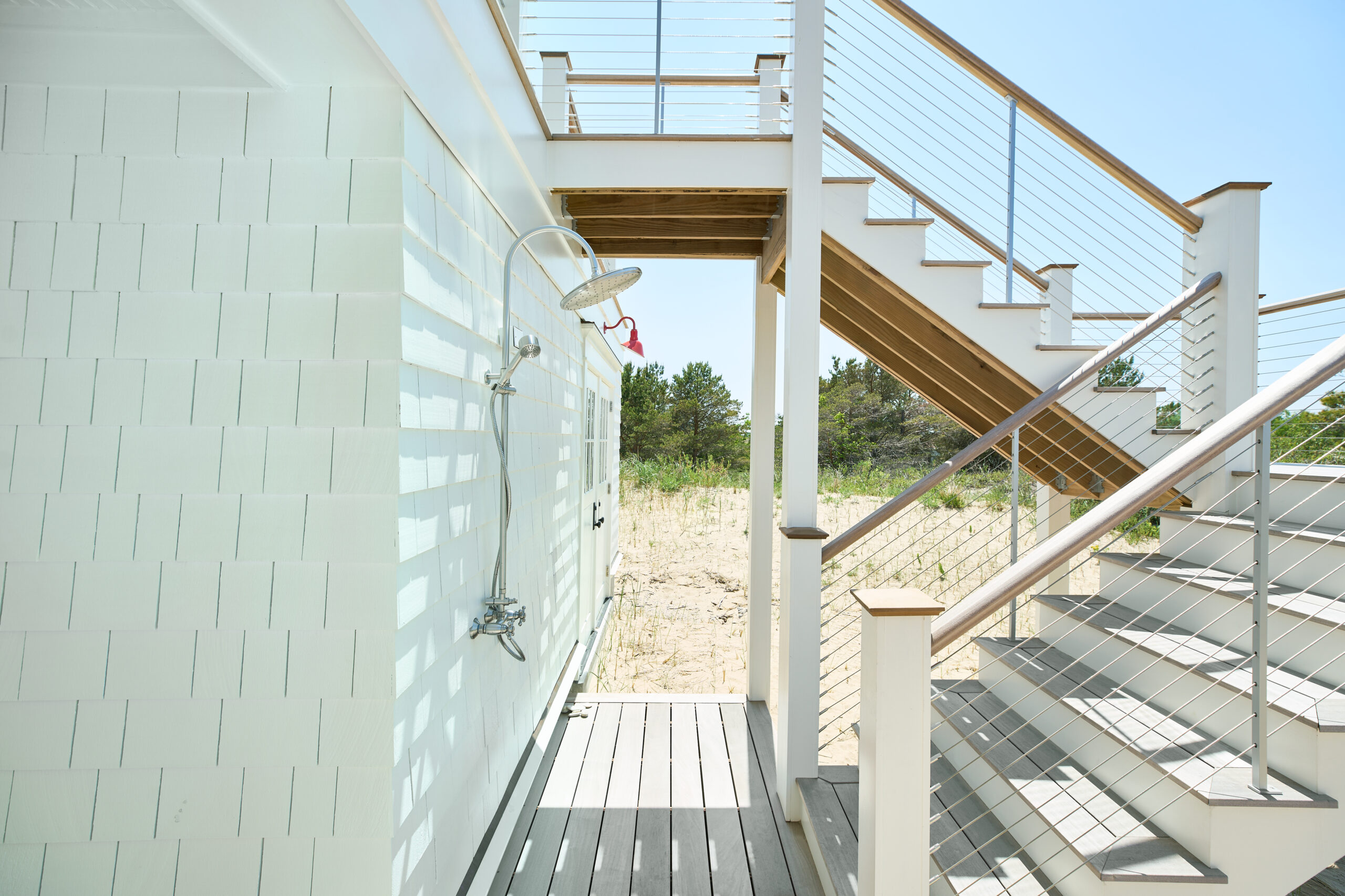 Outdoor beach shower installed on the side of a white shingle-style house beneath a staircase, with sand and dunes in the background.