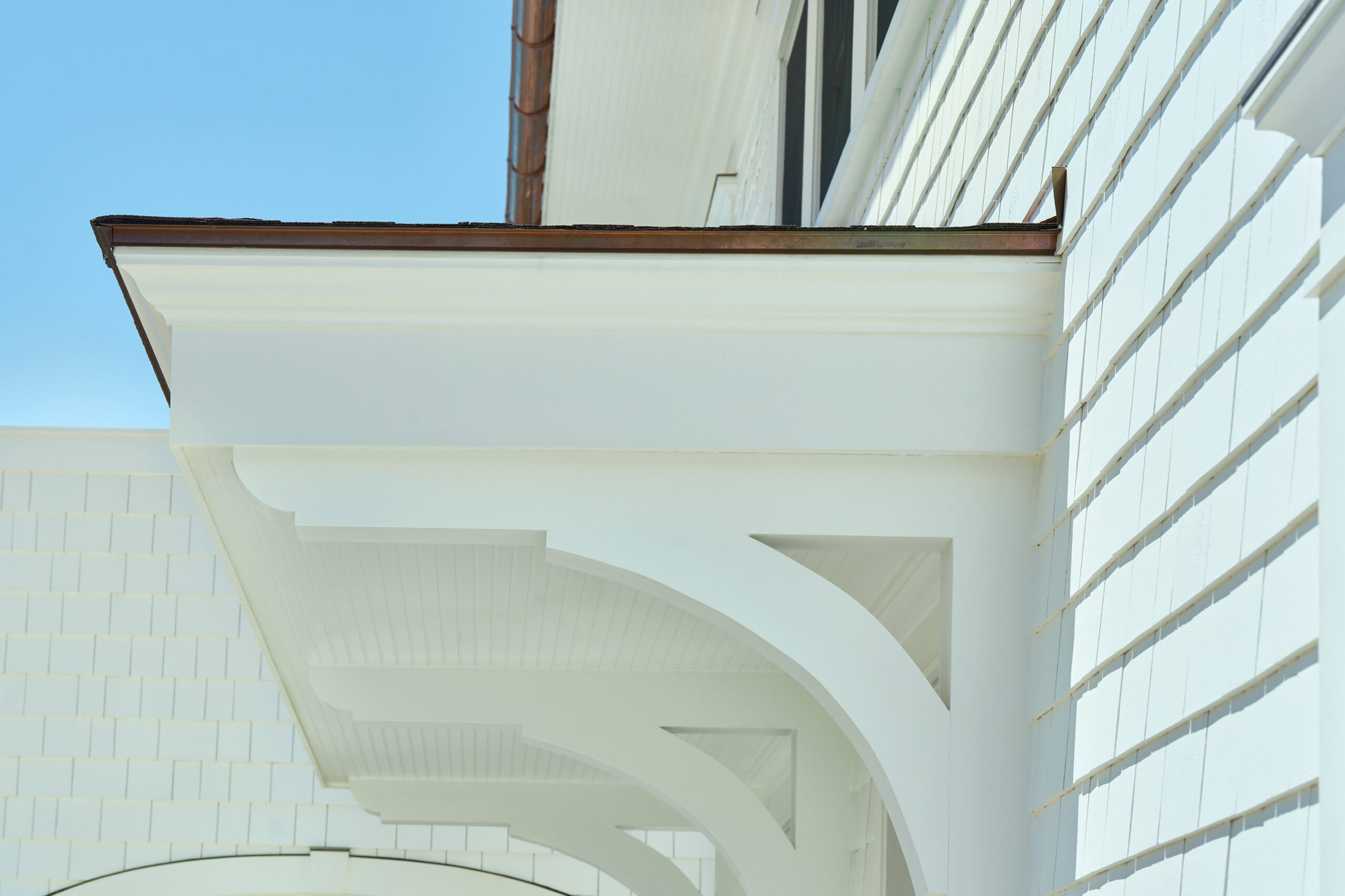Close-up view of white architectural corbels and trim detailing beneath the eaves of a coastal-style home, with beadboard paneling and shingle siding.