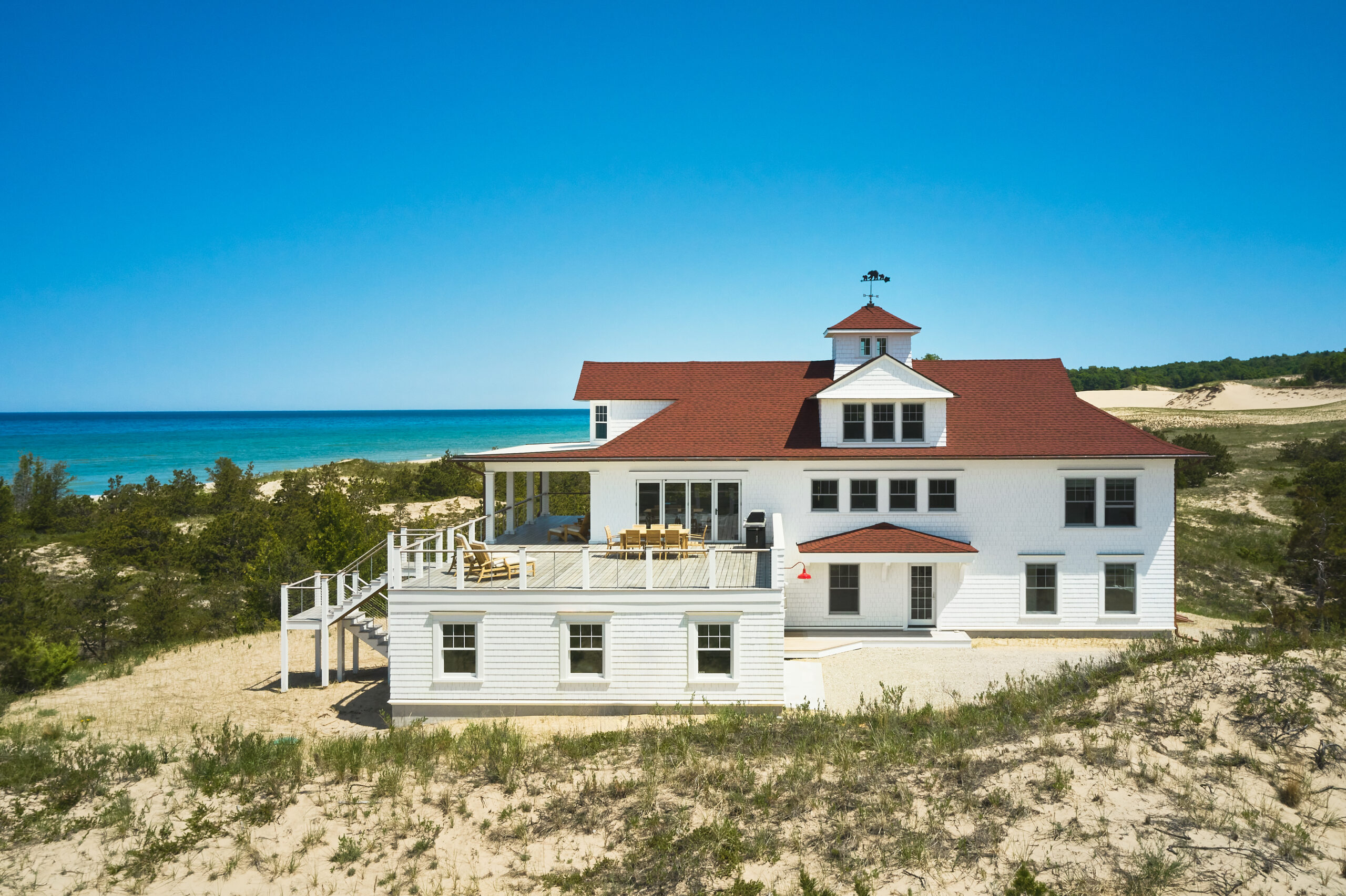 A white cottage with red shingled roofs and a central cupola, perched atop sandy Lake Michigan dunes with the shoreline and blue water visible in the background.