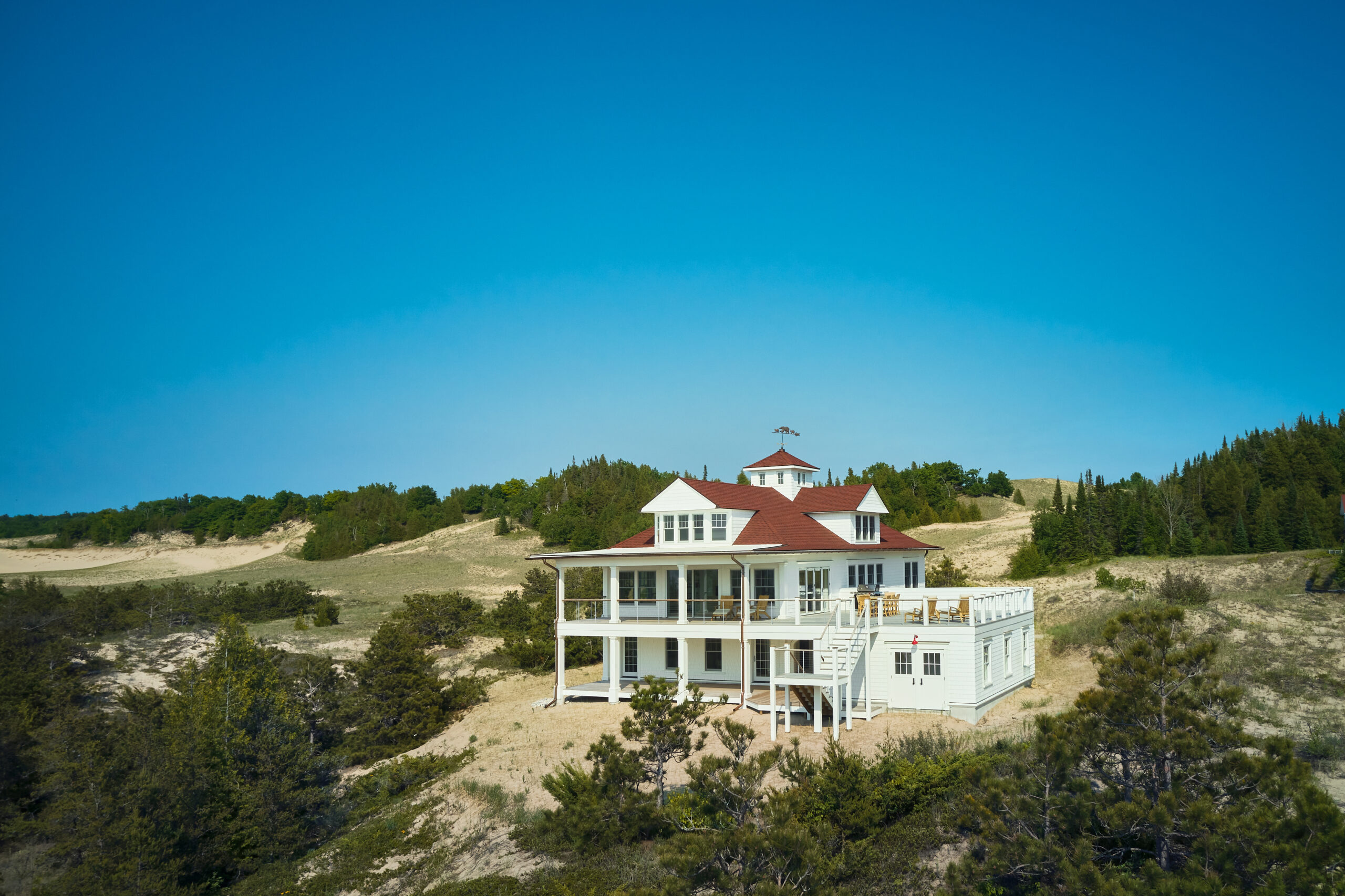 White cottage with red shingled roof nestled among sand dunes and pines under a clear blue sky, designed to resemble the historic Point Betsie Lighthouse.