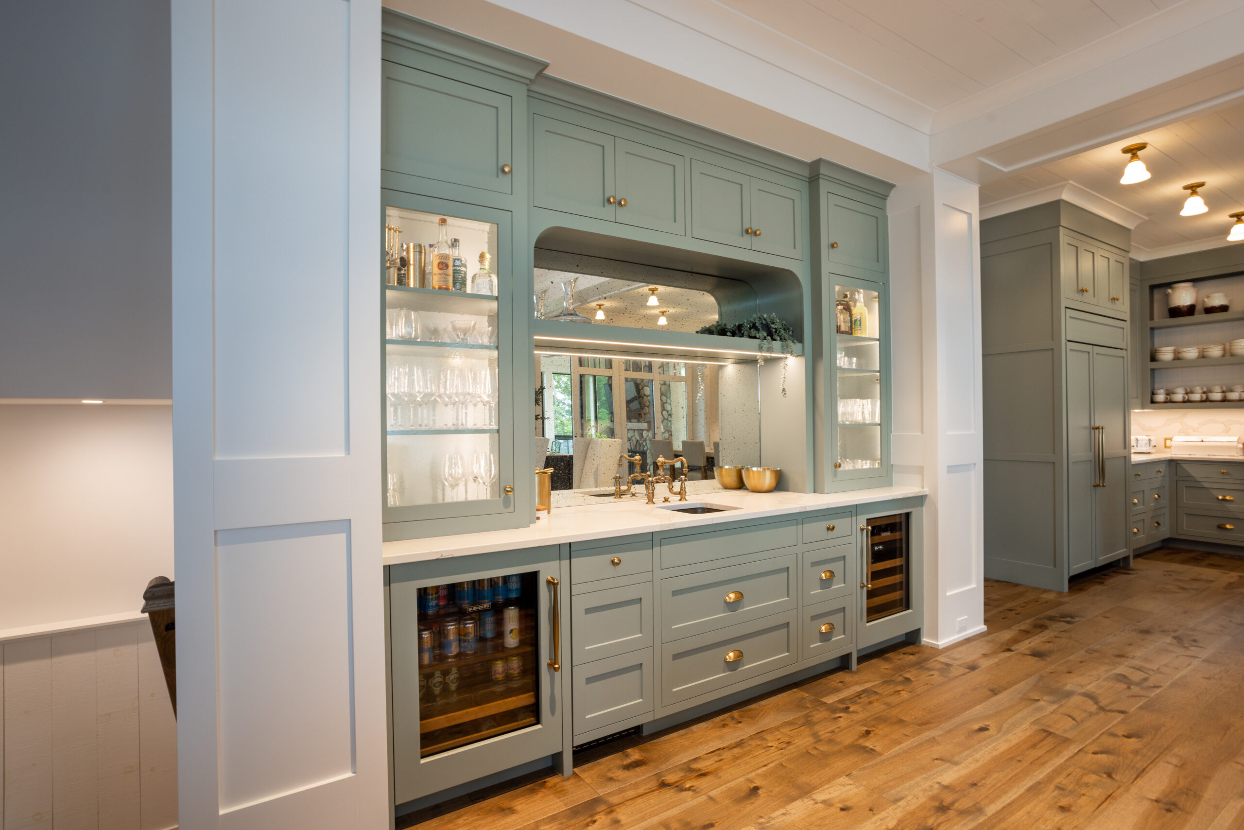 Built-in home bar with sage green cabinetry, glass shelving, and brass hardware.