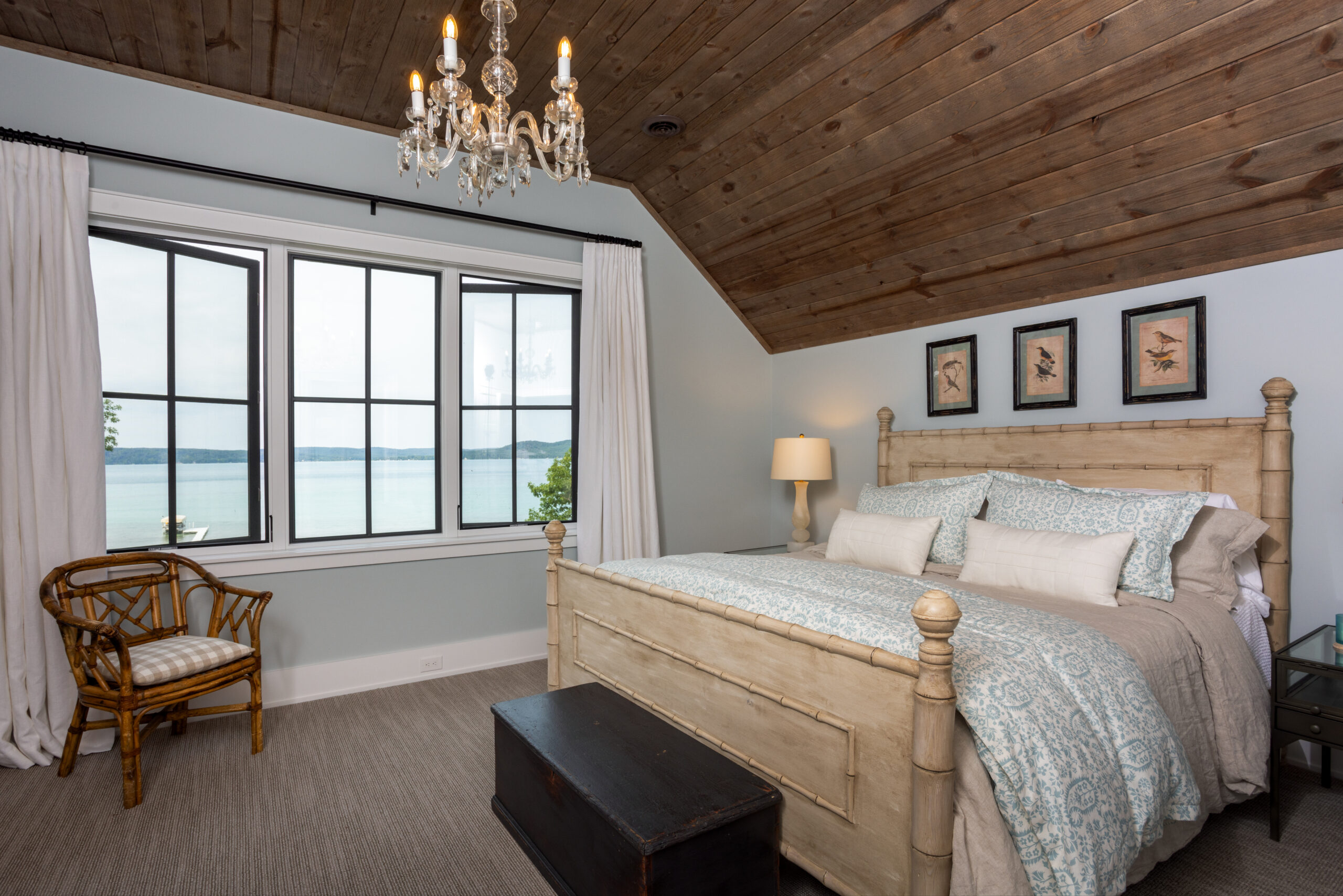 Cozy lakefront bedroom with vaulted wood ceiling, chandelier, and large picture window overlooking the water.