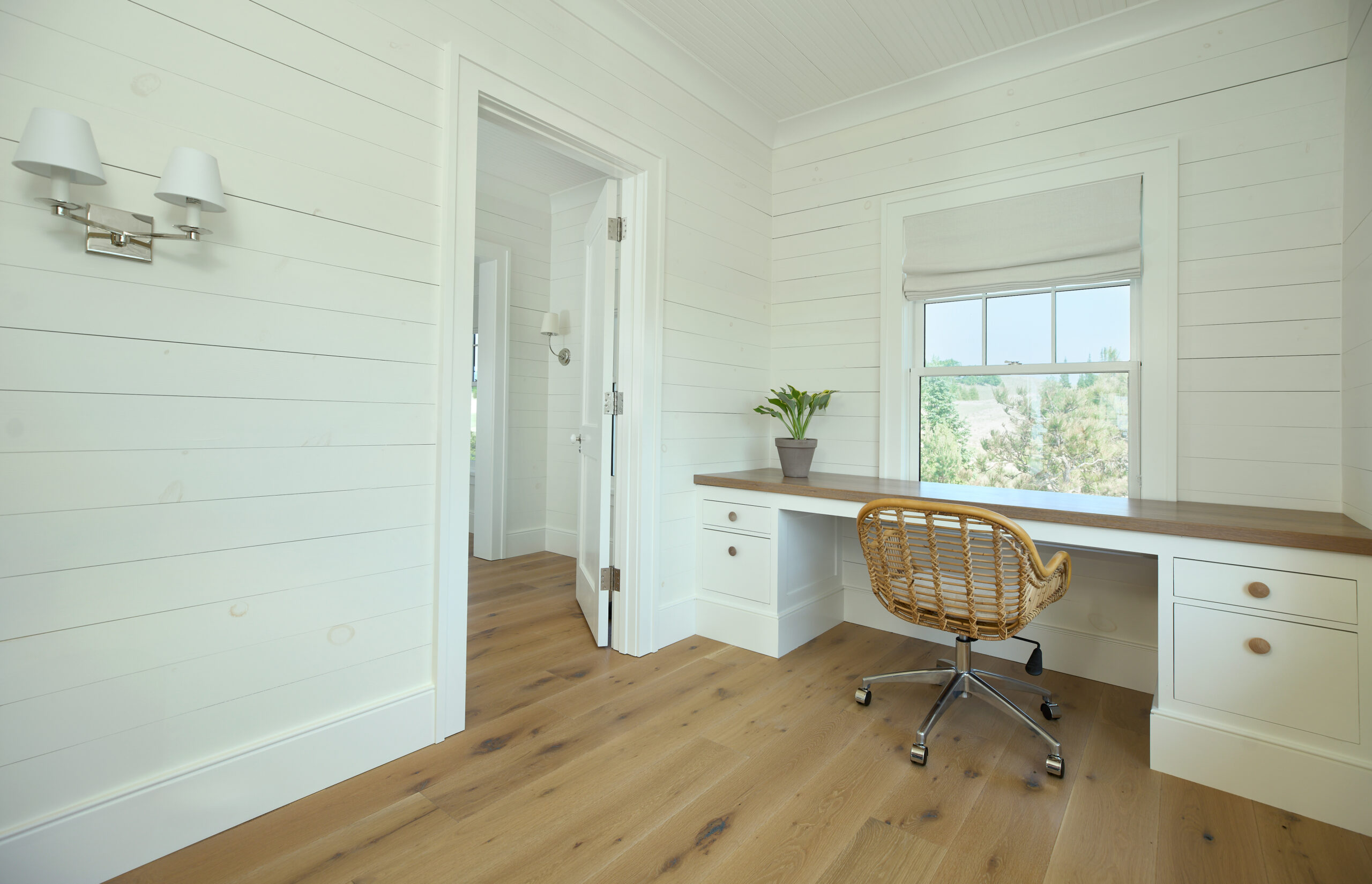 Built-in white desk with wood top, rattan office chair, and window overlooking trees.