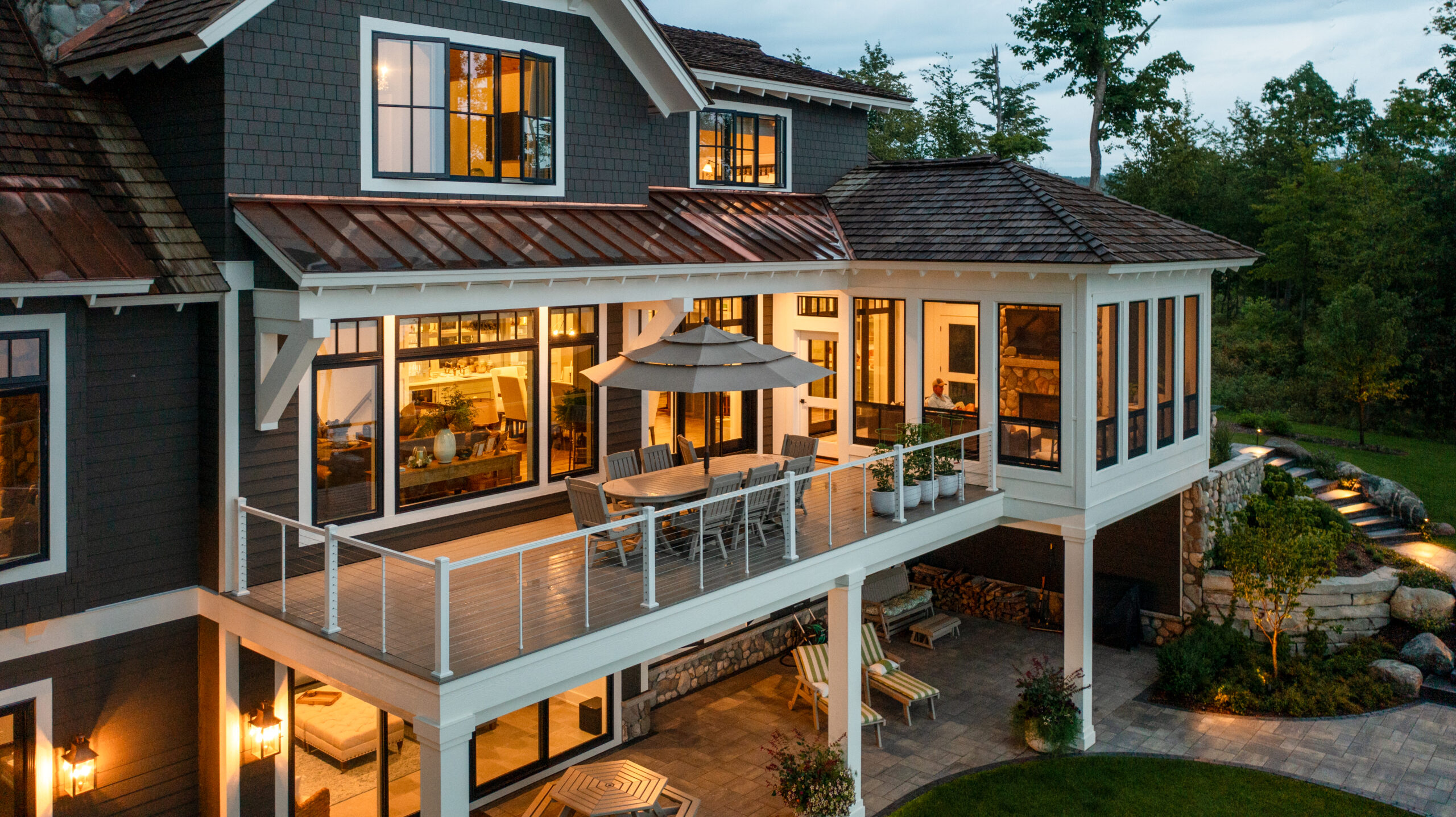 Two-story lake house with upper deck dining area, screened-in porch, and lower patio lounge, all warmly lit at dusk.