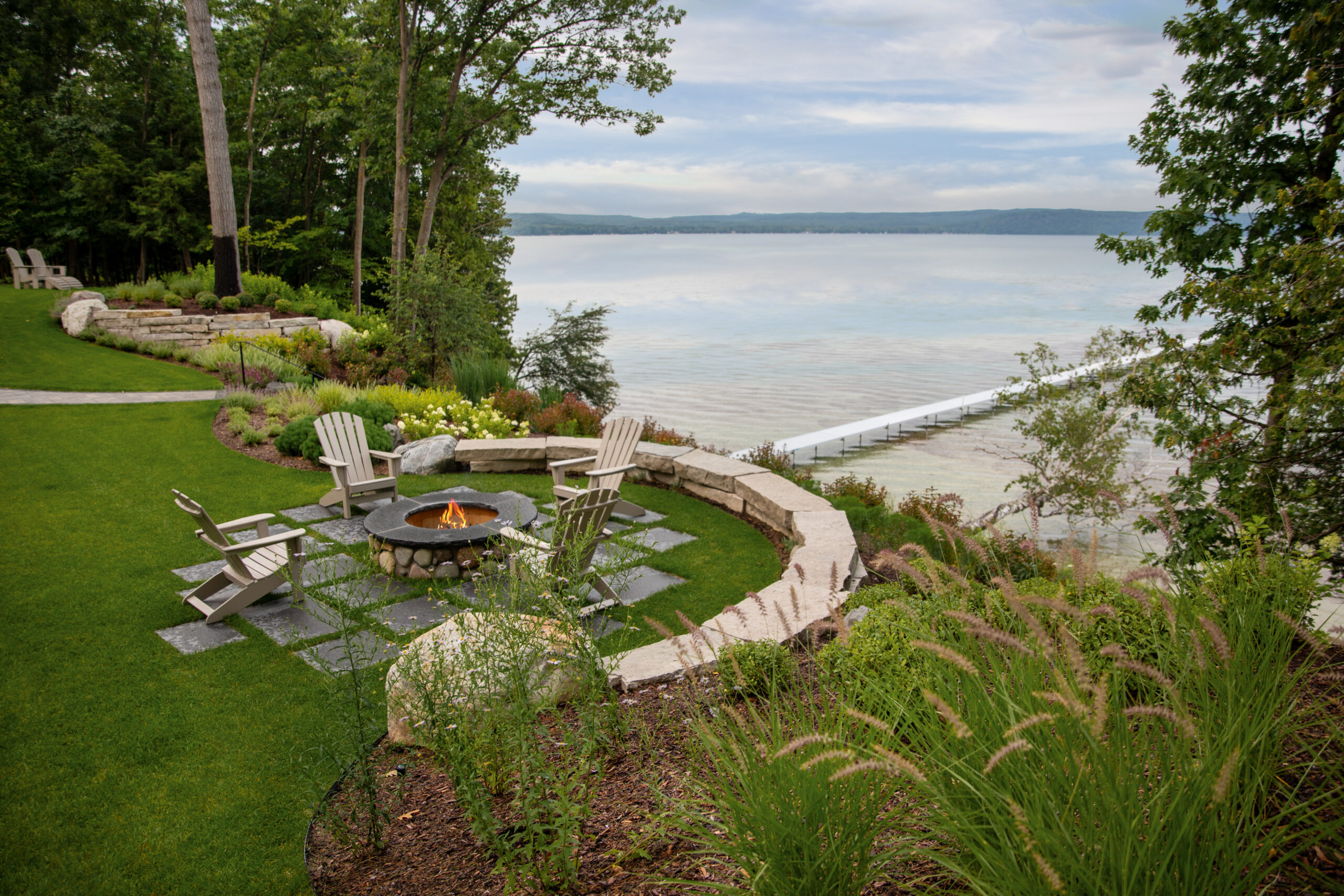 Lakeside fire pit surrounded by Adirondack chairs and lush landscaping overlooking a calm shoreline and dock.