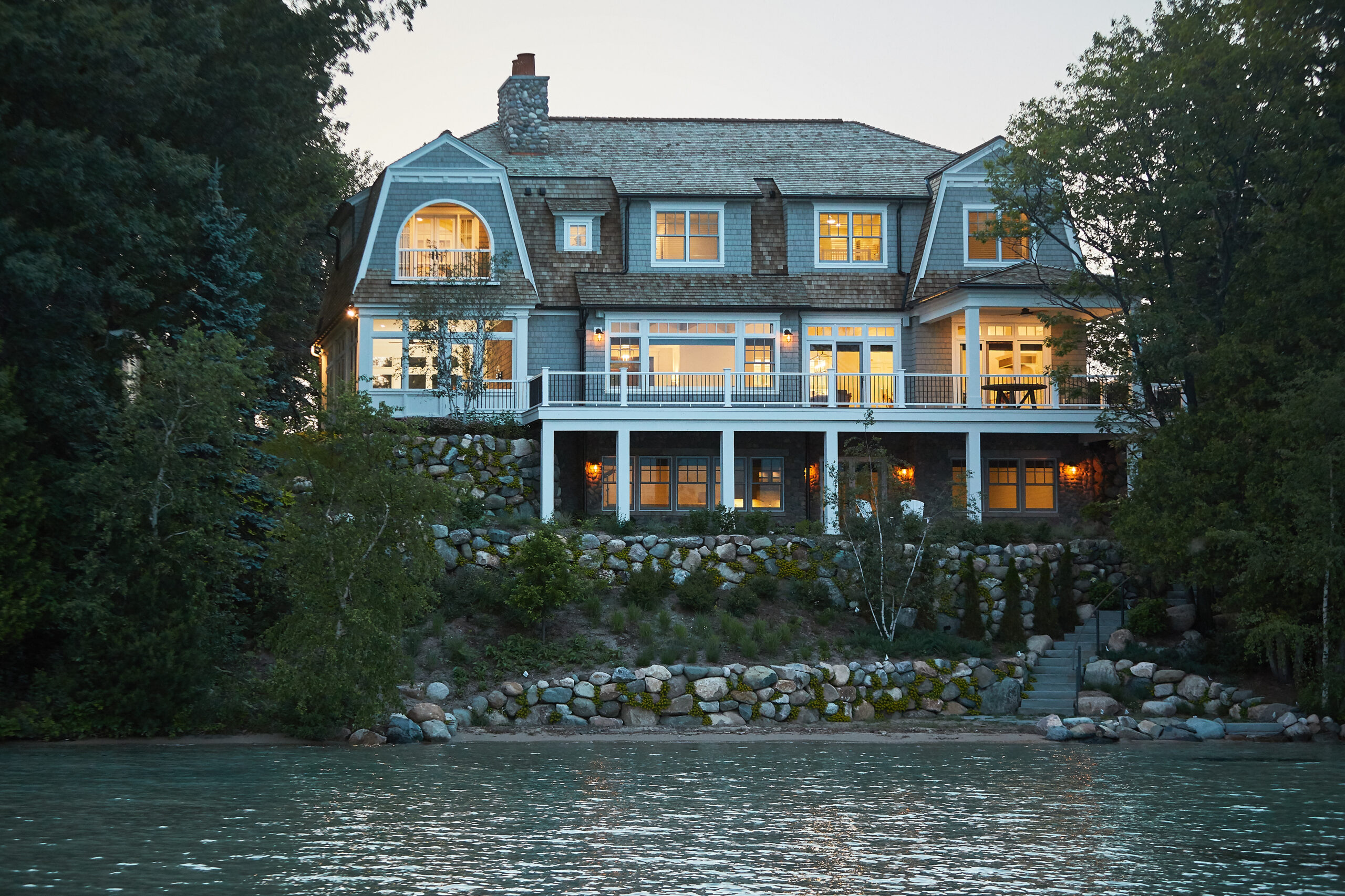 Lakeside home exterior at dusk with illuminated windows and stone shoreline