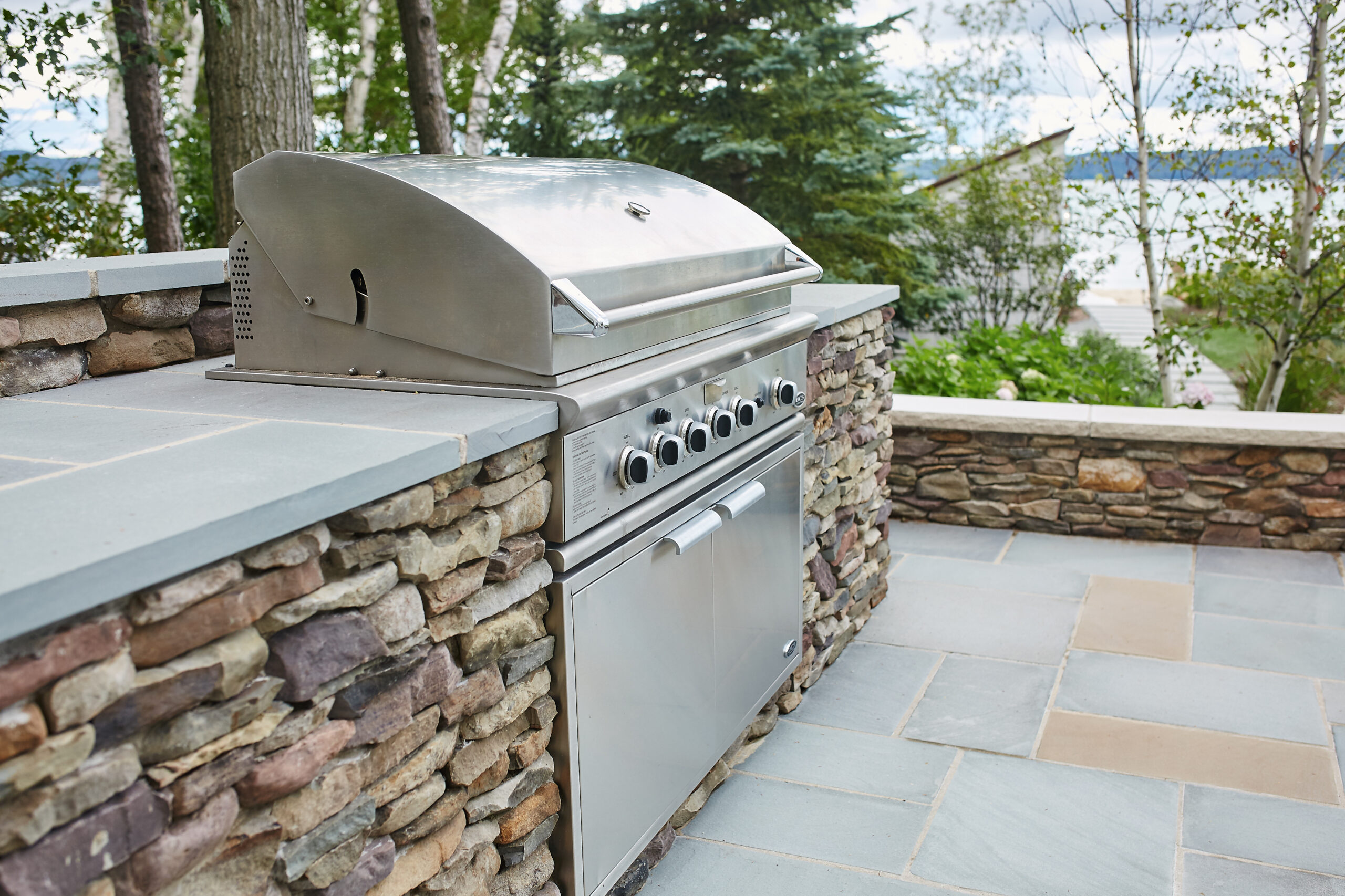 Built-in stainless steel grill set into a stone outdoor kitchen with lake views