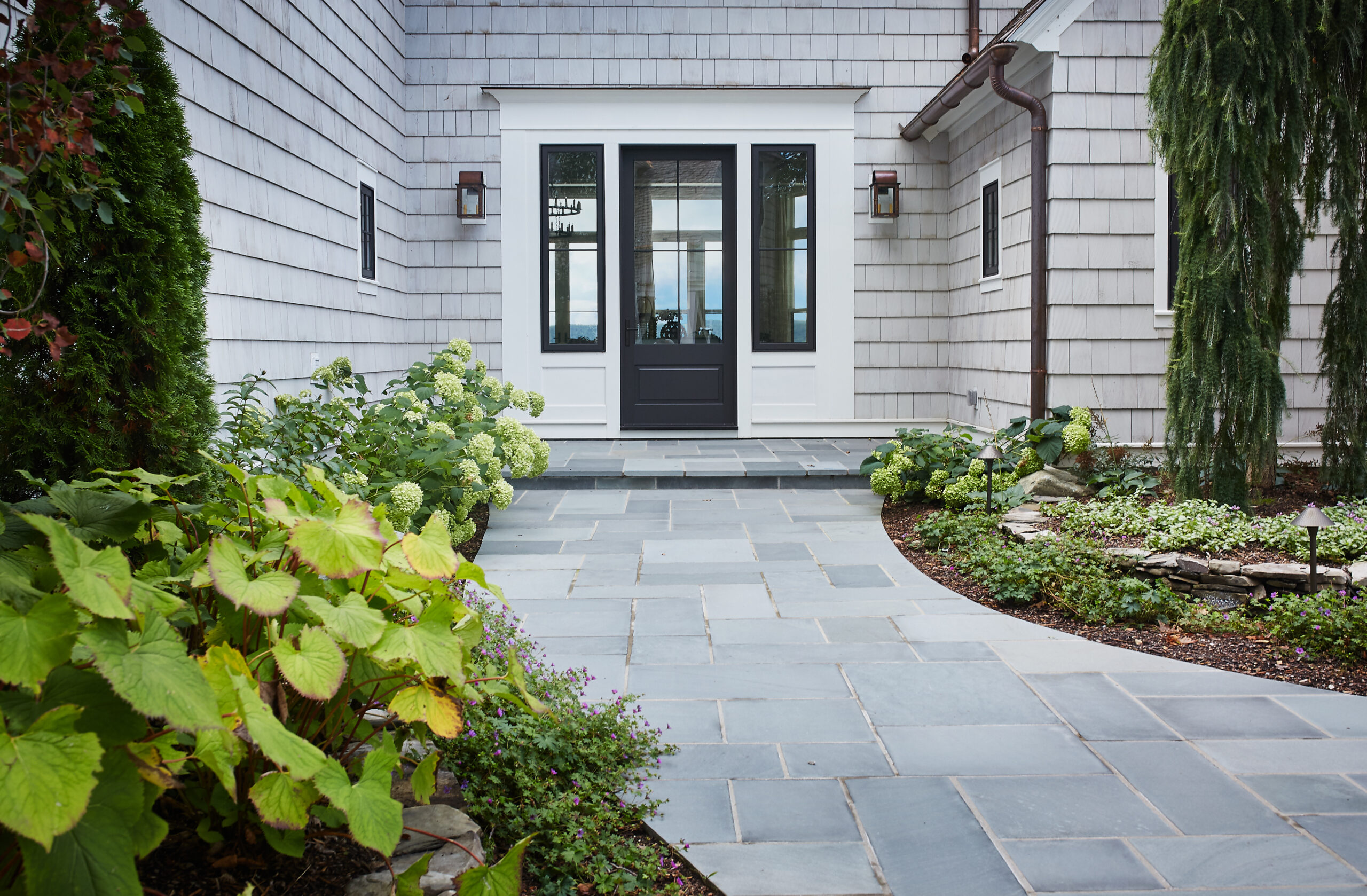 Stone walkway leading to a black front door framed by white trim and lush landscaping