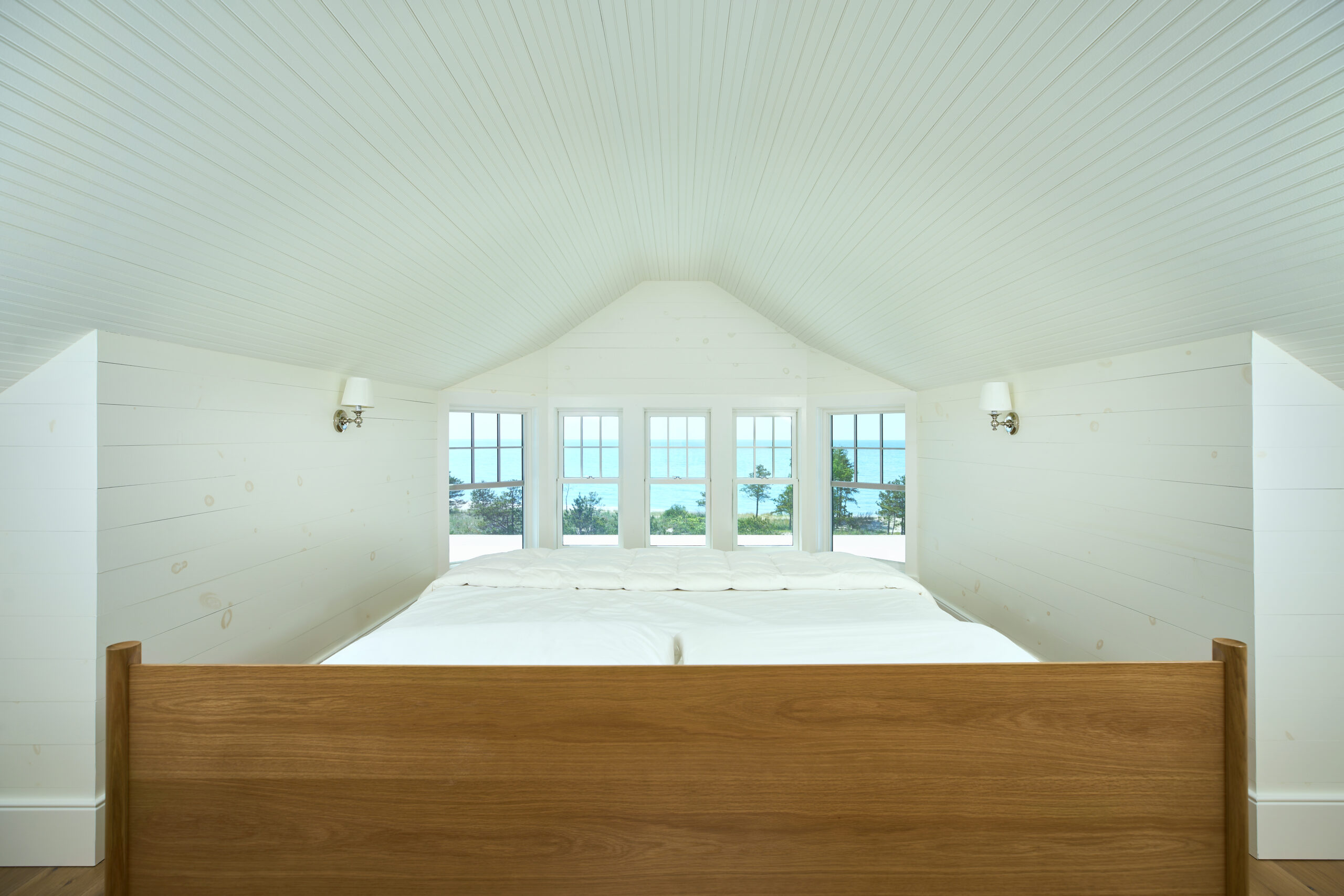 View from a cozy white-paneled bedroom with a peaked ceiling, featuring a warm wood bed frame and expansive windows overlooking Lake Michigan.