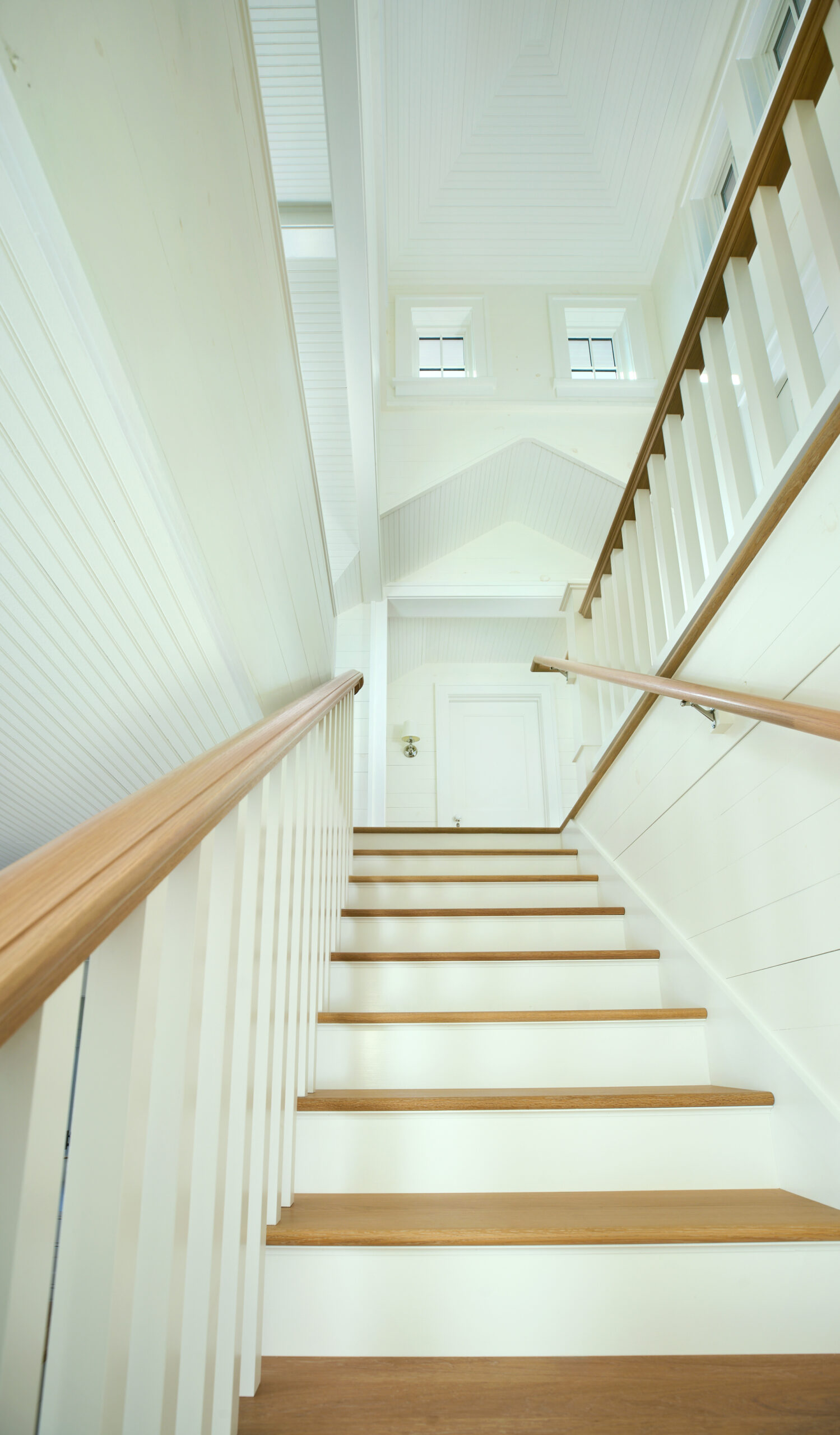Bright white stairwell with natural wood treads and railing, leading up to clerestory windows beneath a vaulted ceiling.