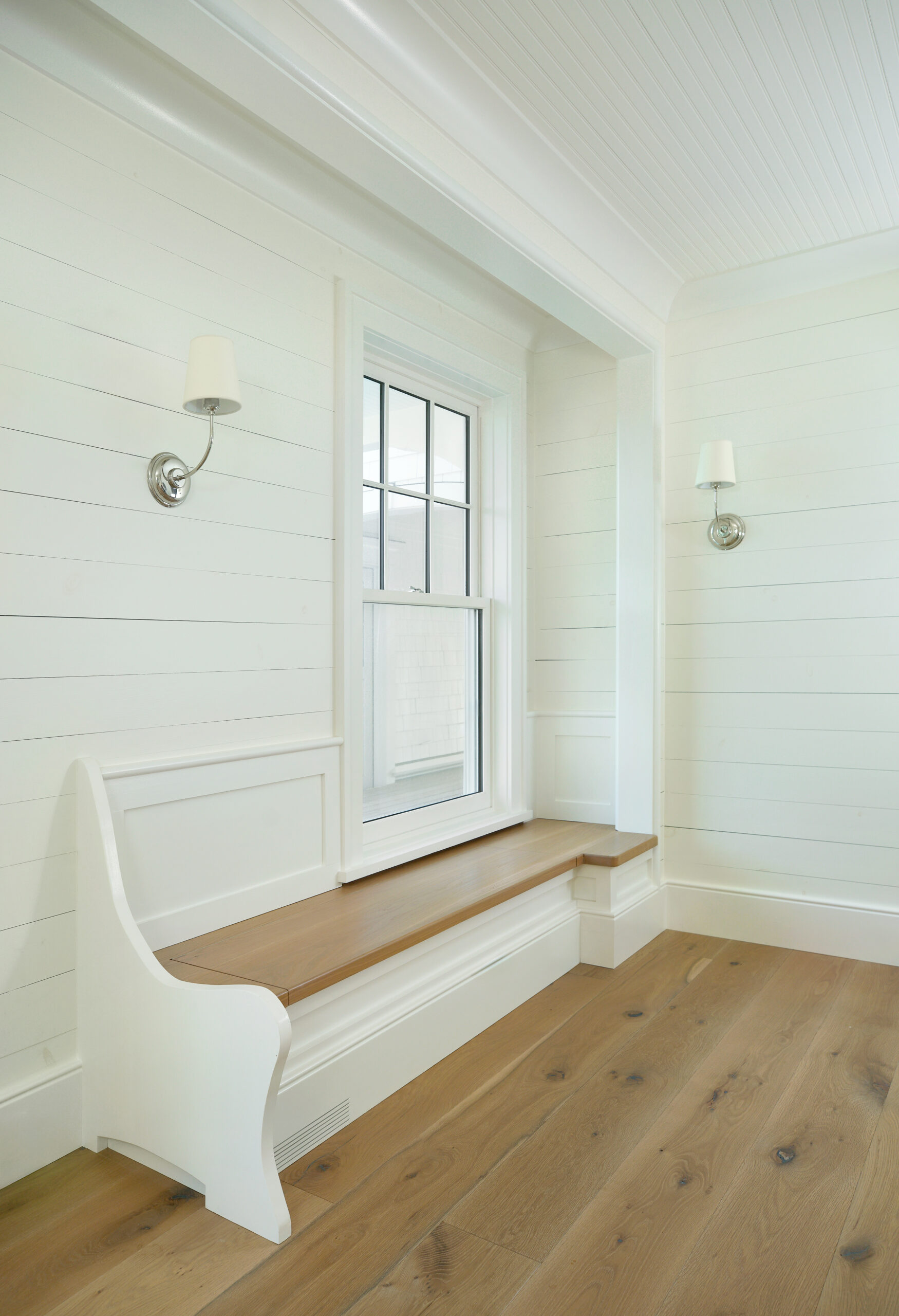 Built-in white wood bench with natural wood seat below a window, framed by shiplap walls and white sconces, atop wide plank wood flooring.