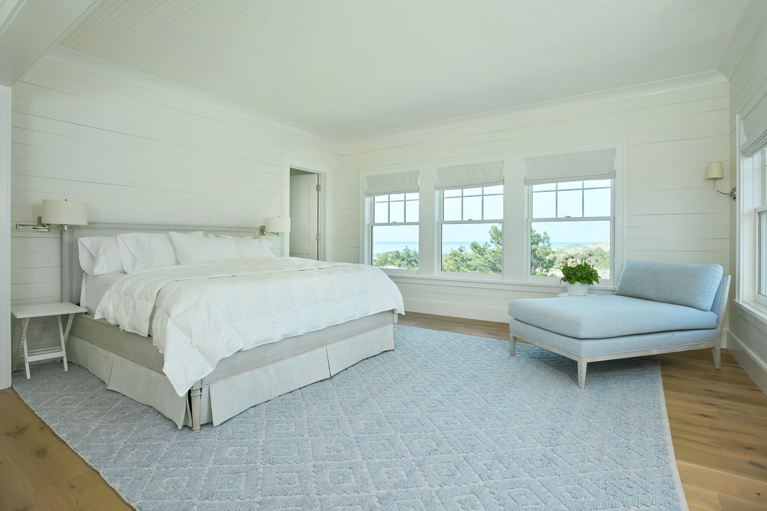 Primary bedroom with large windows, light wood flooring, a white bed, and a blue chaise lounge overlooking Lake Michigan.