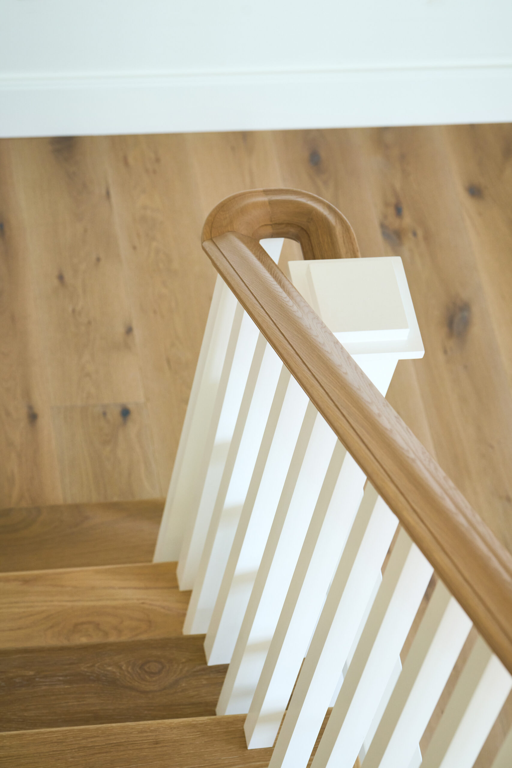 Detail view of a staircase with white balusters and a smoothly curved, natural wood handrail atop warm-toned wood steps and flooring.