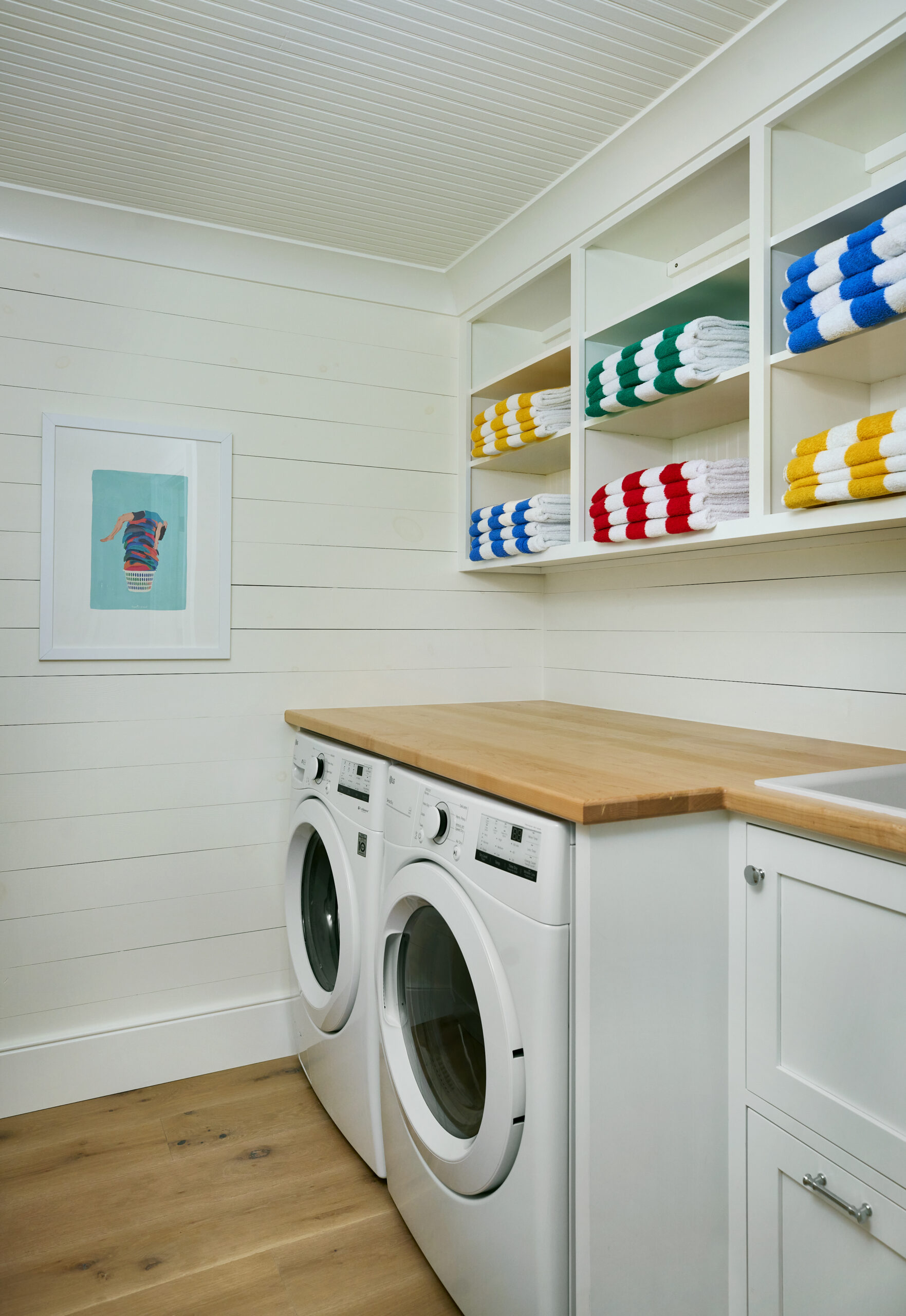 Laundry room with white shiplap walls, stacked white washer and dryer, butcher block countertop, and colorful striped towels on open shelves.