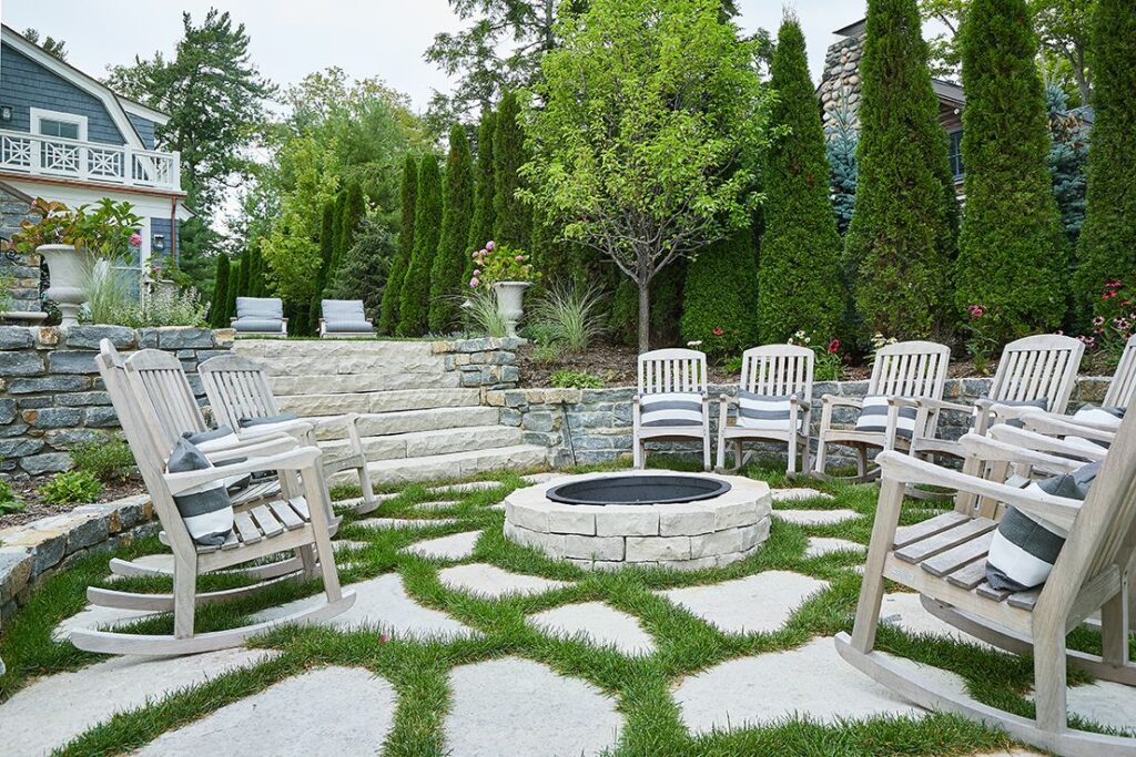 Outdoor fire pit area with a circular stone fire feature surrounded by wooden chairs, set within a landscaped yard with stone retaining walls and greenery.