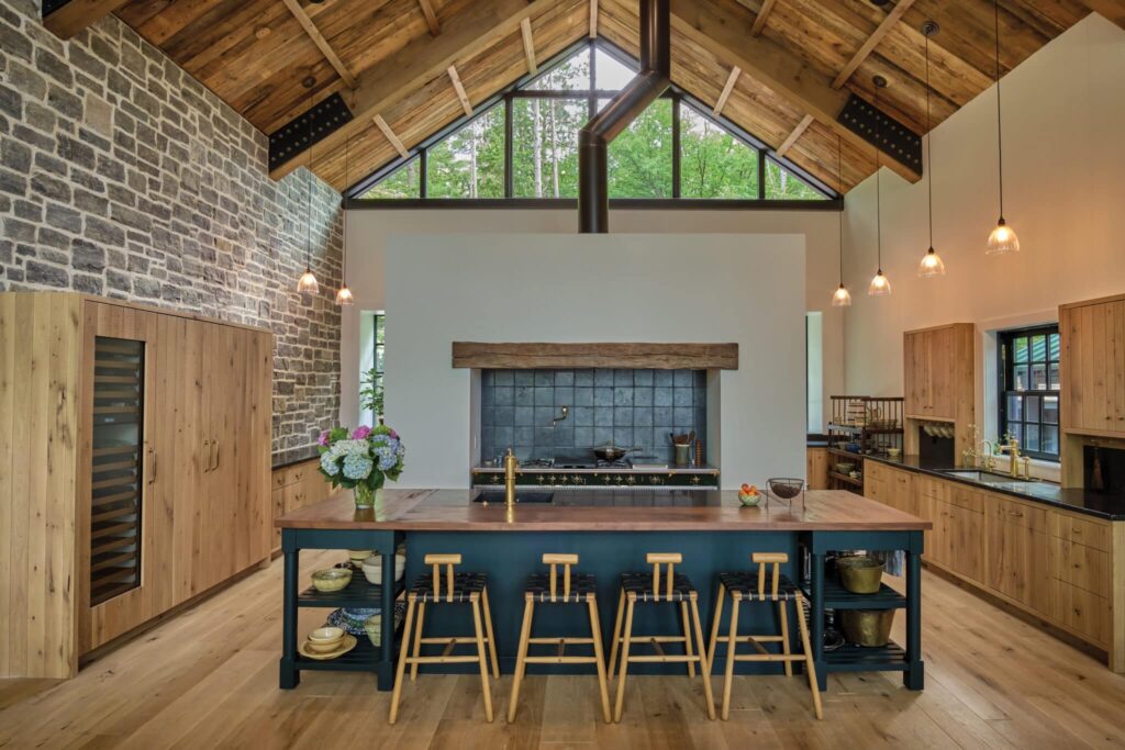 Spacious kitchen with a large island, wood cabinetry, vaulted wood ceiling, stone accent wall, and expansive windows overlooking trees.