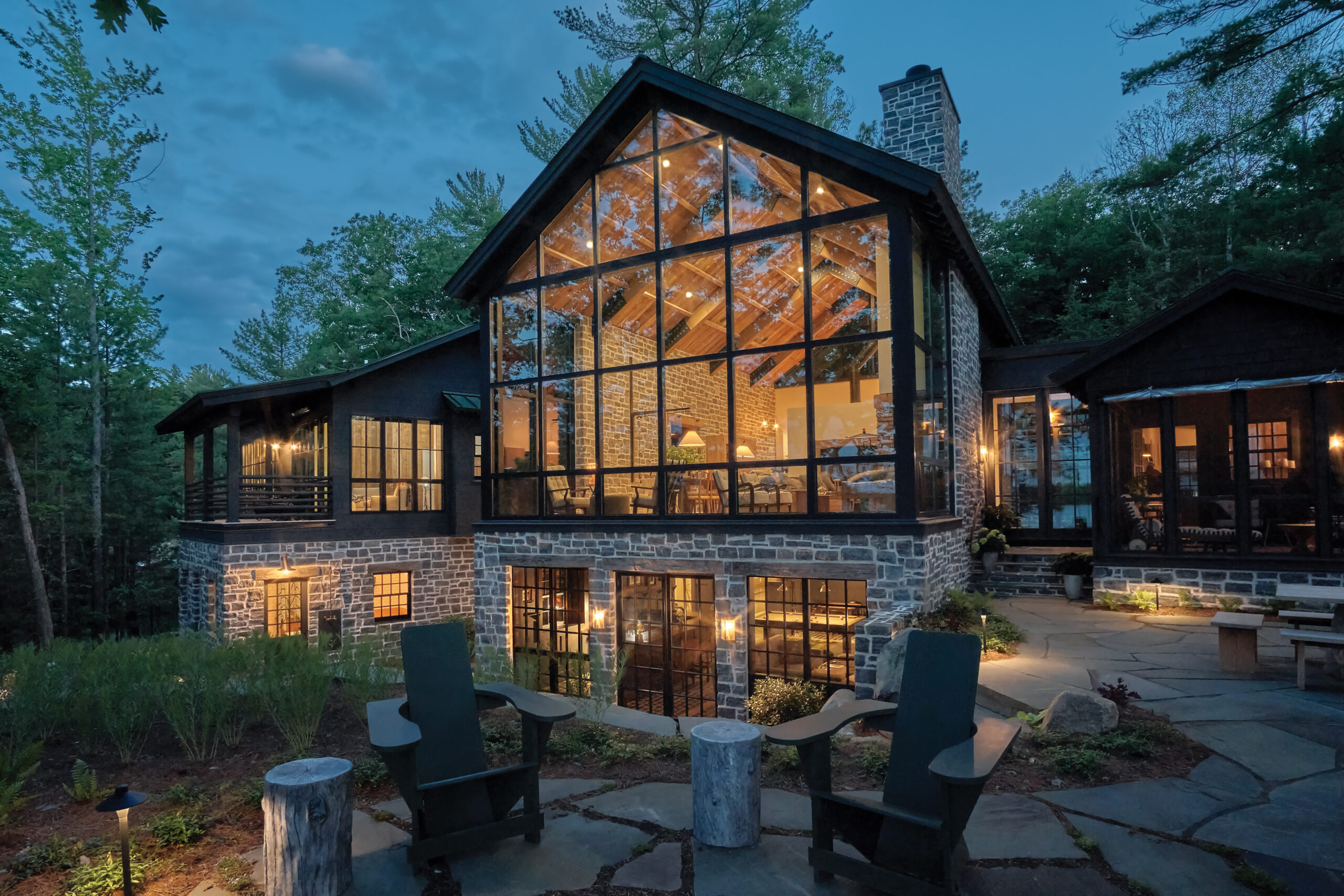 Exterior view of a rustic lakefront home at dusk with large glass windows, reclaimed stonework, and warm interior lighting, surrounded by forest.