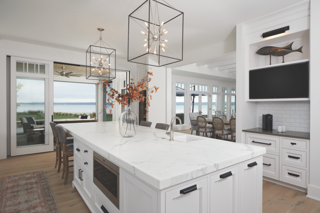 White kitchen with large marble island, pendant lighting, and open view to dining area and lakeside patio through glass doors.