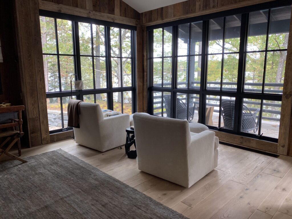 Corner seating area with two upholstered chairs beside large black-framed windows overlooking trees and a lake.