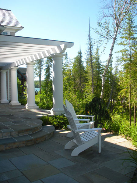 Stone patio with white pergola, columns, and Adirondack chairs overlooking wooded landscape