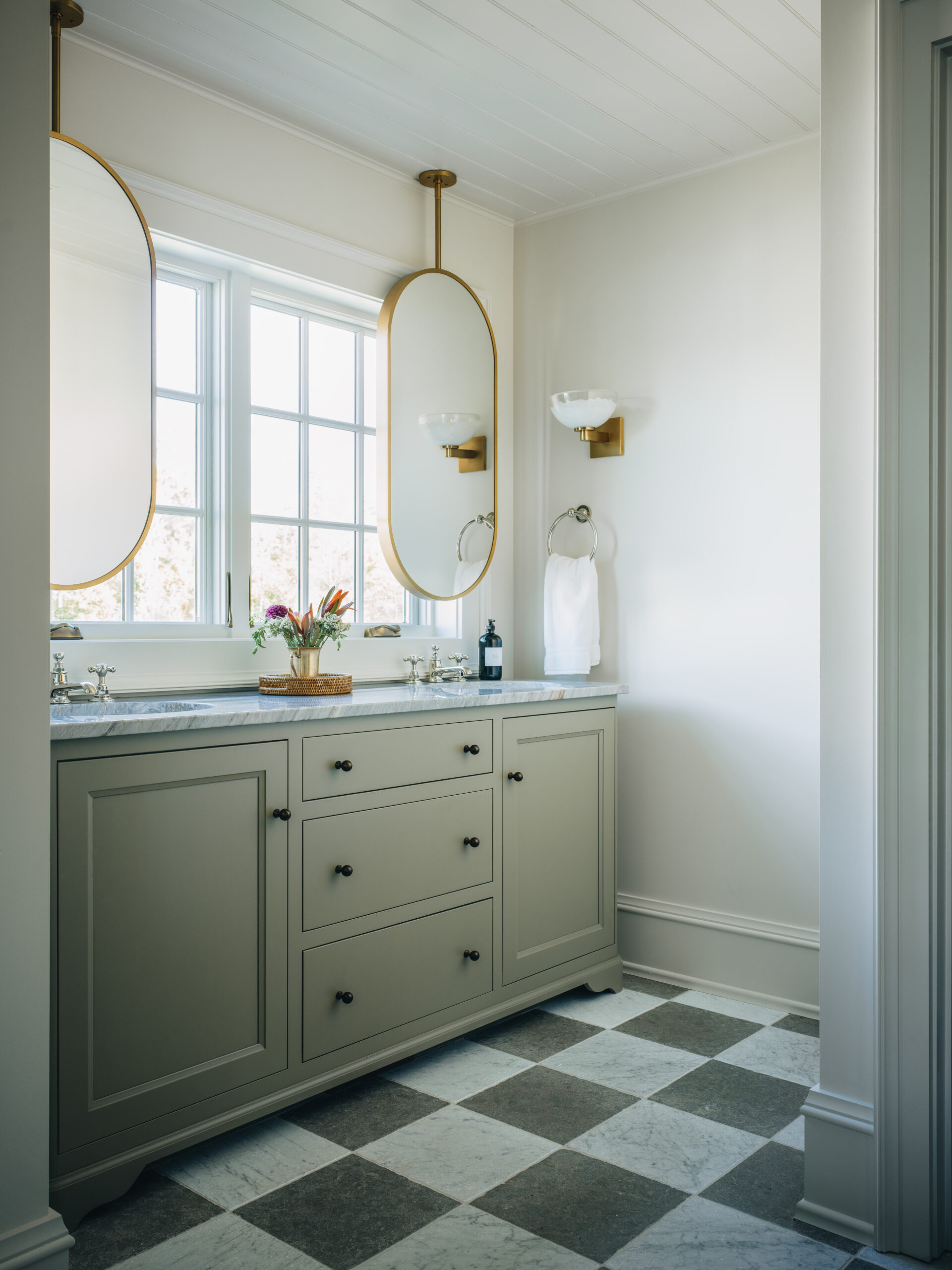 Elegant guest cottage bathroom with checkered marble flooring, dual vanity with sage cabinetry, and suspended oval mirrors in front of a large window.