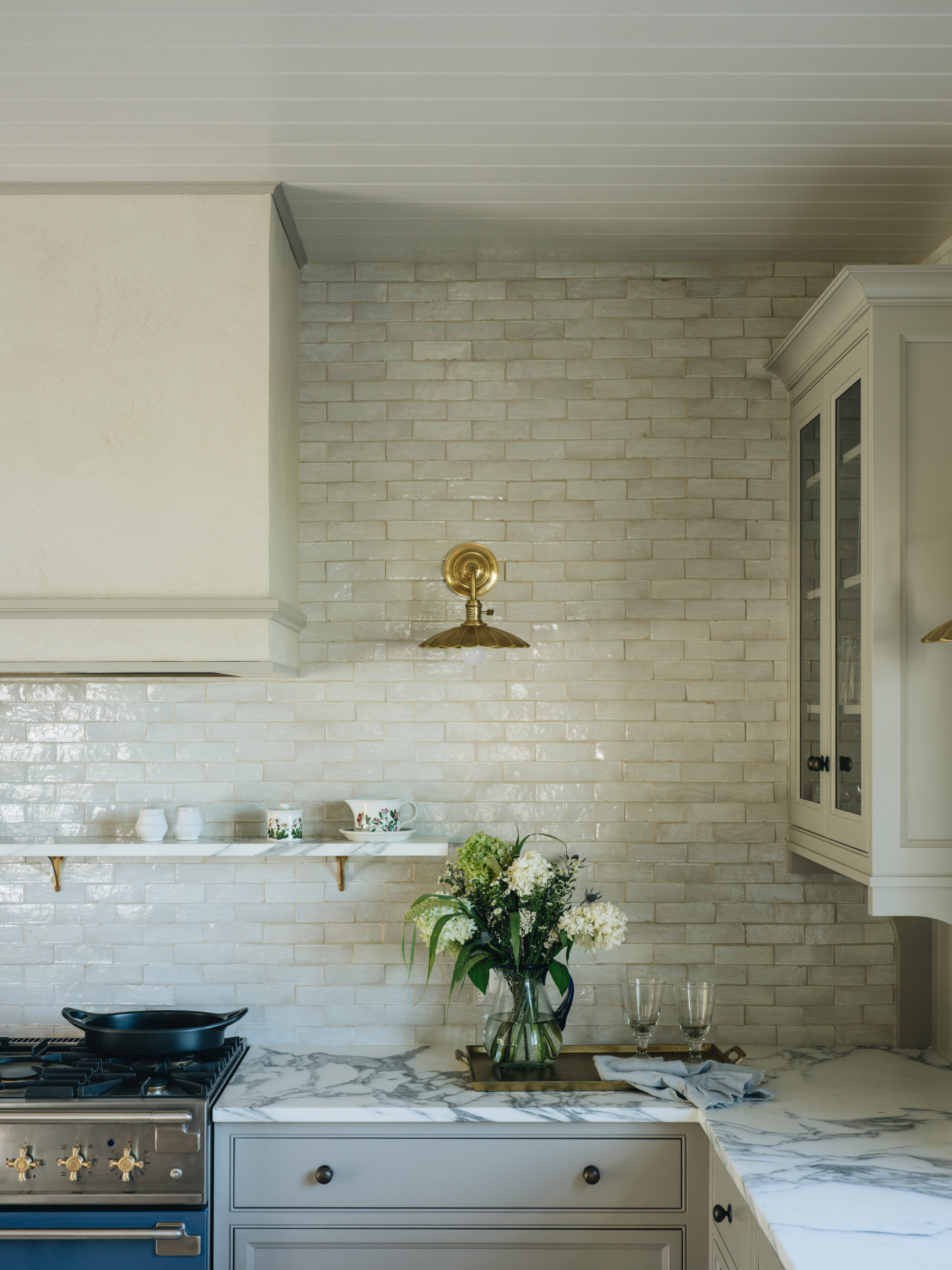 Marble countertop kitchen with soft gray cabinetry, brass accents, a blue range, and white brick backsplash in a guest cottage.