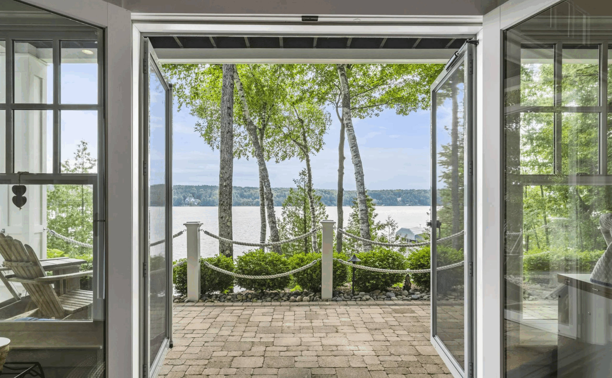 Open glass doors framing a lakeside patio with stone pavers, trees, and water view beyond
