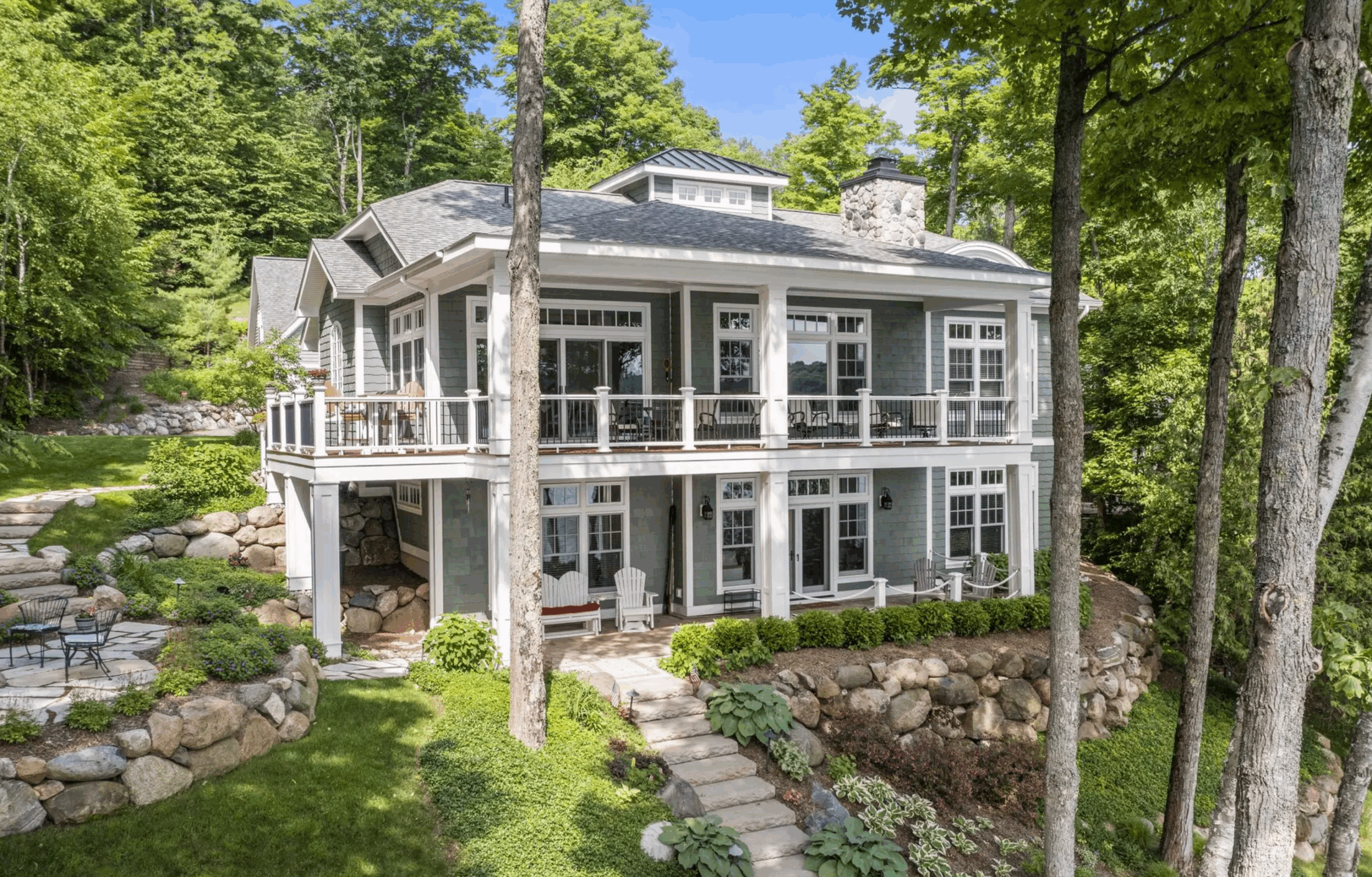 Two-story lakeside cottage with wraparound porch, stone terraces, and wooded hillside setting
